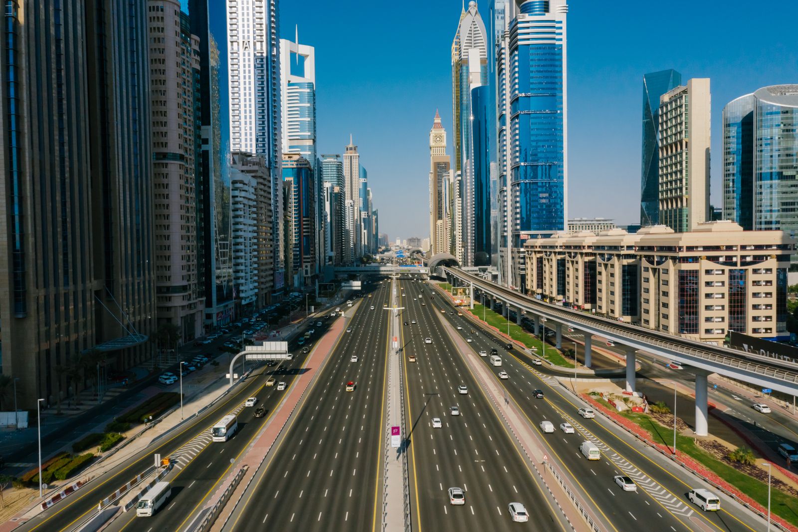 Aerial view of a wide multi-lane Dubai highway with a Metro viaduct and glass skyscrapers either side