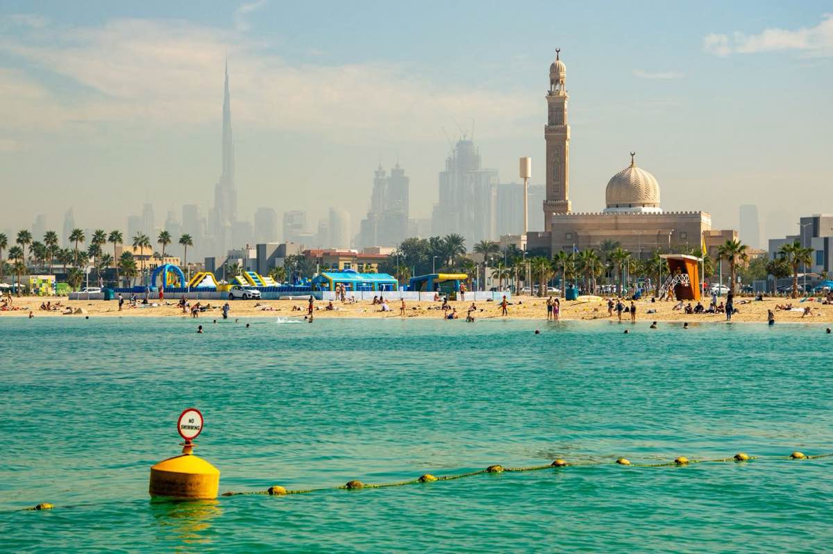 Dubai beach in winter with clear turquoise water, palm trees, a mosque and the Burj Khalifa skyline behind